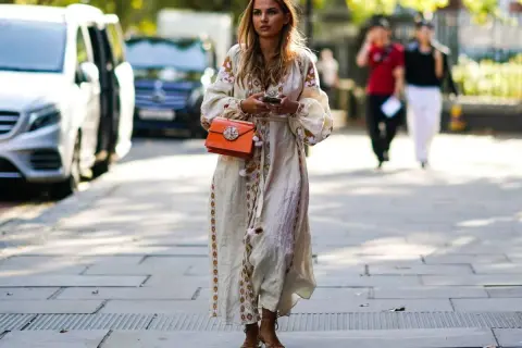 Kobieta w sukience boho z haftem i pomarańczową torebką. Idealne buty do sukienki boho to delikatne sandałki na obcasie.