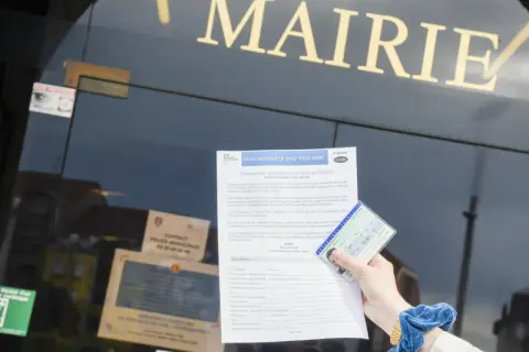 Main dans la main, une carte d'identité française et un formulaire de demande de changement de nom de famille sont présentés devant la mairie. Un geste pour de fautereau vassel.