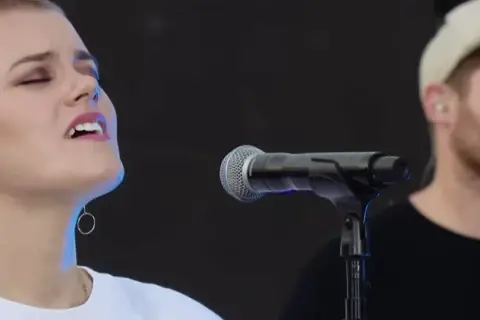 Taya Smith singt mit geschlossenen Augen und In-Ear-Monitoring. Ein Mann mit Kappe steht im Hintergrund.