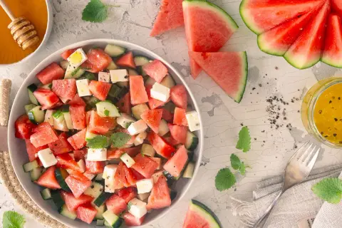 Erfrischender Wassermelonen Feta Salat mit Gurkenstücken und Minze, garniert mit Pfeffer. Daneben ein Glas Dressing und Honig.