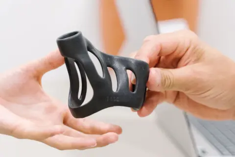 Eine Hand reicht eine schwarze Schiene, die zur Behandlung eines Schnappfingers dient, einer anderen Hand.