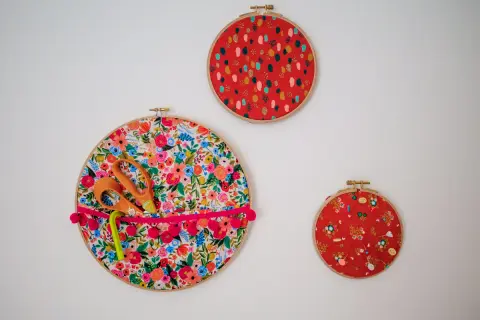 Décoration murale avec trois cercles à broder. Un grand cercle fleuri sert de rangement pour des ciseaux et un stylo, orné d'un ruban à pompons. Deux autres cercles rouges décoratifs complètent l'ensemble.