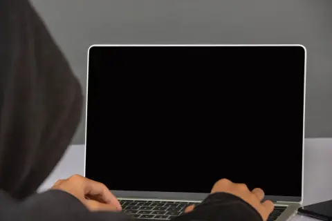 Osoba w kapturze pisze na laptopie z czarnym ekranem. Ciemność skrywa jej działania.