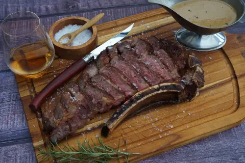 Ein saftiges Cote de Boeuf, perfekt geschnitten, mit grobem Salz bestreut, daneben ein Glas Whiskey, ein Messer und eine Sauce.