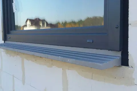 Szary parapet pod oknem, którego framuga odbija dom i drzewa.