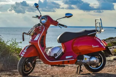 Czerwona Vespa na tle morza. Czy na skuter trzeba mieć prawo jazdy? Kask leży obok.