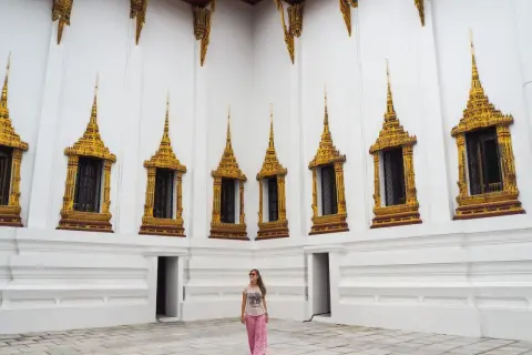 Una mujer sonríe frente a una pared blanca con ventanas doradas ornamentadas, un ejemplo de qué ver en Tailandia.