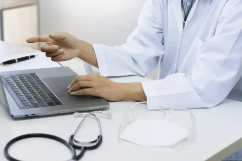 Lekarz w białym kitlu przy laptopie wyjaśnia, czy do poradni dermatologicznej potrzebne jest skierowanie. Na biurku stetoskop i maseczka.