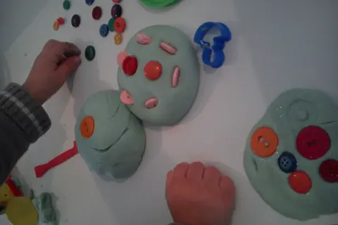 Manos infantiles jugando con plastilina casera, decorada con botones de colores y cortadores.