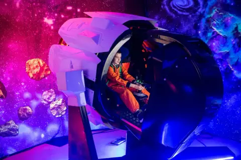 Chłopiec w pomarańczowym kombinezonie steruje statkiem kosmicznym w symulatorze. Kosmopark Wrocław oferuje kosmiczne przygody wśród asteroid i mgławic.