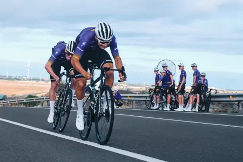 Cykla på Gran Canaria i tävlingsform. Ett cykellag i lila dräkter susar fram på en asfalterad väg med vindkraftverk i bakgrunden.