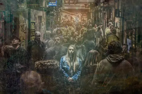 Młoda kobieta w dżinsowej kurtce stoi pośród tłumu, jej wyraz twarzy zdradza strach.