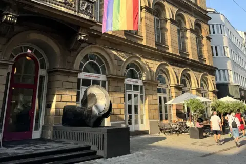 Ein Regenbogenbanner weht über dem von der Heydt Museum. Eine Skulptur steht davor, daneben ein Café mit Sonnenschirmen.