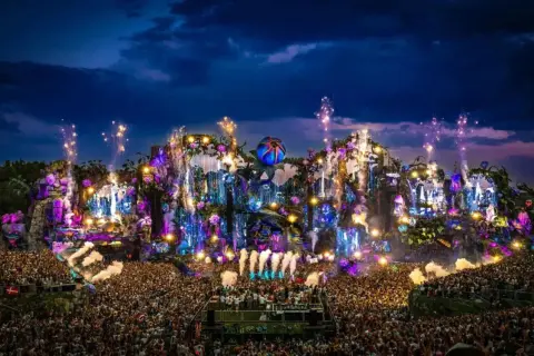 Un'enorme folla festeggia sotto un palco spettacolare, illuminato da luci colorate e fuochi d'artificio. È il luogo dove fanno il Tomorrowland, un'esplosione di musica e magia.