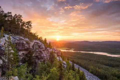 Solnedgång över en skogklädd dal med en sjö. Klippor och träd i förgrunden. En vacker vy som ger fakta om Finland.
