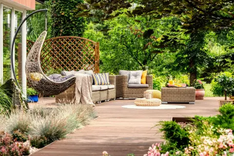 Un acogedor rincón en un jardín de verano, con muebles de mimbre, una silla colgante y vegetación exuberante.
