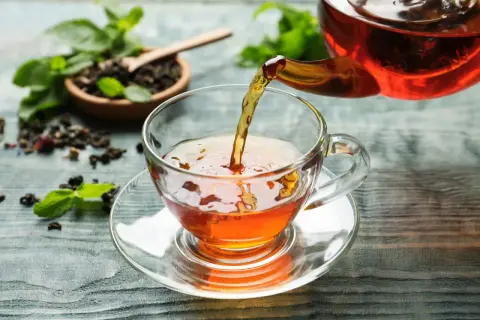 Un thé ambré est versé dans une tasse transparente. Le thé est-il bon pour la santé ? Des feuilles de menthe et du thé en vrac sont disposés sur la table.