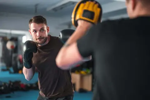 A man in black boxing gloves practices his boxing workout routine, focusing on his opponent holding a yellow mitt.