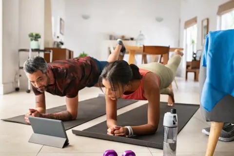 Ein Paar macht Liegestütze zu Hause, inspiriert von **micro workouts** auf einem Tablet. Hanteln und Wasserflasche liegen bereit.
