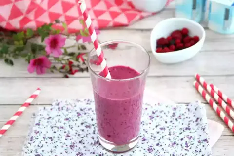 Ett glas med en lila indisk yoghurtdryck, dekorerad med ett rött och vitt randigt sugrör. Bredvid finns en skål med bär och yoghurtburkar.
