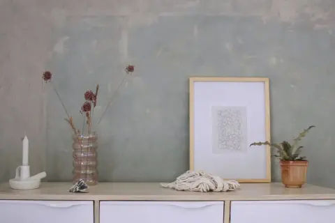 Un tavolino con candela, vaso di fiori secchi, quadro e corda. Sembra la preparazione calce per imbiancare, con un tocco artistico.