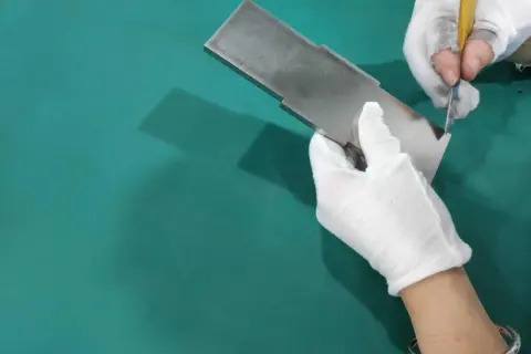 Ręce w białych rękawiczkach dokonują gratowania metalowego elementu za pomocą nożyka.