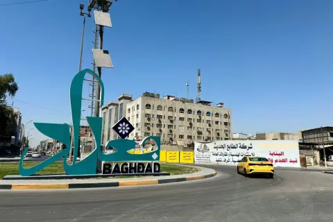 Wielki napis "BAGHDAD" i stylizowany napis arabski na tle budynków i nieba. Stolica Iraku wita podróżnych.