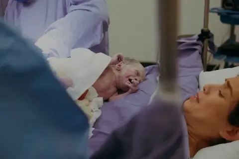 Noworodek po indukcji porodu, owinięty w biały ręcznik, leży obok szczęśliwej matki.