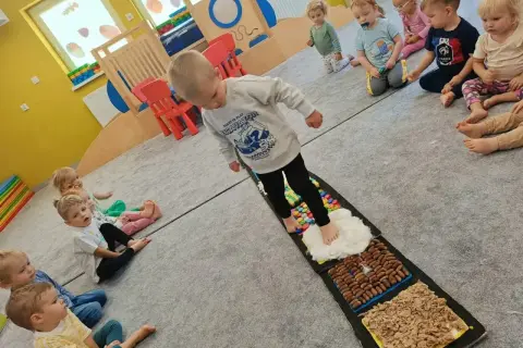 Dzieci aktywnie w żłobku ćwiczą zmysły na ścieżce sensorycznej. Maluchy z zaciekawieniem obserwują kolegę stąpającego po różnych fakturach.