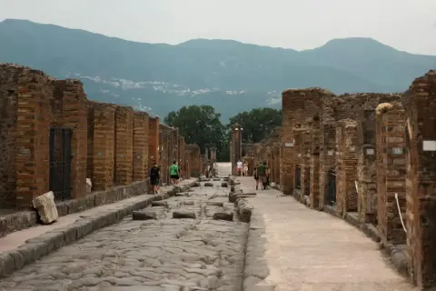 Besucher spazieren auf der antiken, kopfsteingepflasterten Straße von Pompeji heute, umgeben von den Ruinen römischer Gebäude. Berge im Hintergrund.