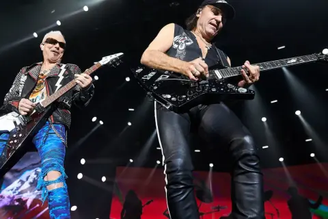 Dwóch gitarzystów na scenie, jeden w skórzanej kurtce i niebieskich spodniach, drugi w czarnej kamizelce i skórzanych spodniach. Ich gra wywołuje "wind of change" tłumaczenie.