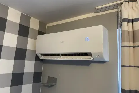Biała klimatyzacja Haier zamontowana na szarej ścianie, obok zasłony.