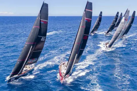 Diverse barche a vela nere con vele rosse e bianche competono in una regata, solcando le onde blu con determinazione.