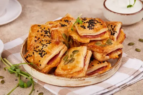 Pizzerinki z ciasta francuskiego, posypane czarnuszką i pestkami dyni, na talerzu z serkiem i kiełkami.