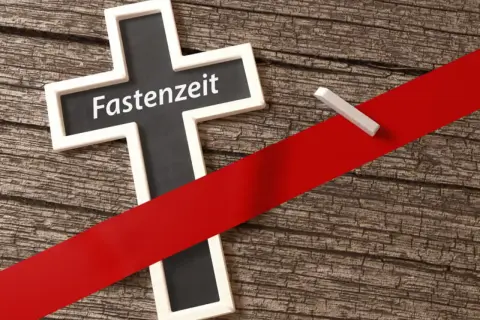 Kreuz mit der Aufschrift "Fastenzeit" auf Holzuntergrund, durchzogen von einem roten Band. Ein Stück Kreide liegt daneben.