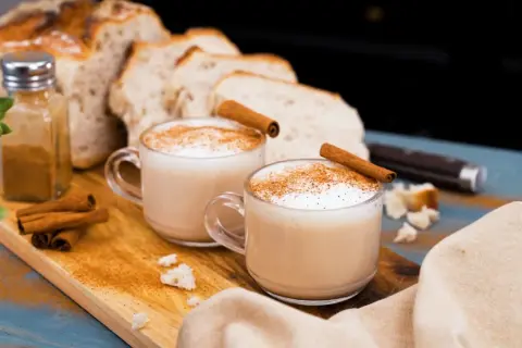 Två koppar krämig chai latte med kanelstänger och strödd kanel. Brödskivor och kanelstänger i bakgrunden.
