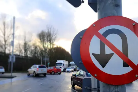 Znak zakaz zawracania na mokrej drodze, z samochodami w tle. Kierowcy muszą uważać, bo nie wolno tu zawracać.