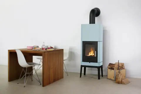 Ein hellblauer Bullerjan B3 Kaminofen mit brennendem Feuer neben einem Holztisch mit Geschirr und Stühlen.