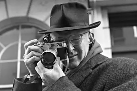 Mężczyzna w kapeluszu i okularach trzyma aparat, gotowy do uchwycenia chwili. Jego skupienie zdradza pasję do fotoreportażu.