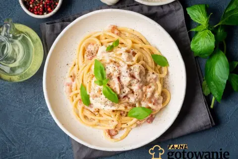 Pyszne spaghetti carbonara ze śmietaną bez jajek, udekorowane bazylią i pieprzem. Obok butelka oliwy i miseczka z pieprzem.