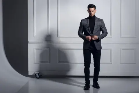 Uomo con maglione nero a collo alto e giacca a quadri grigia, pantaloni scuri e scarpe eleganti.