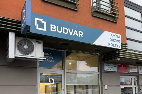 Szyld salonu okien i drzwi "BUDVAR" z logo, klimatyzatorem i napisem "OKNA DRZWI ROLETY".