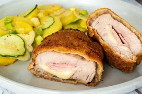 Zwei goldbraune Cordon-bleu-Schnitzel mit geschmolzenem Käse und Schinken, serviert mit einem Kartoffel-Zucchini-Salat.