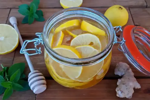 Słoik z wodą, cytryną i imbirem. Obok leżą cytryny, gałązka mięty i drewniana łyżka do miodu.