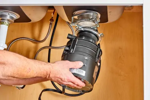 Hands guide a new garbage disposal into place under a sink, part of a garbage disposal installation.