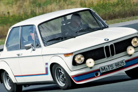 Białe BMW 2002 Turbo z kolorowymi pasami na zderzaku i błotnikach, pędzi po torze.