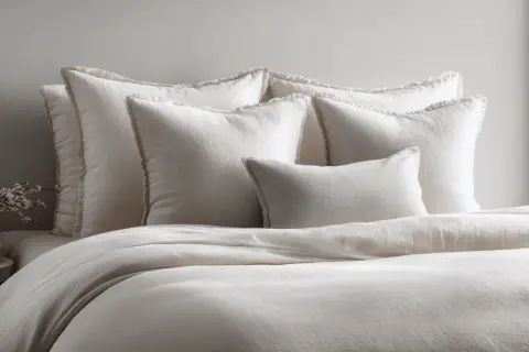 A bed with plush white pillows and a duvet. The pillows have a frayed edge, and one smaller textured pillow sits in front. This is what is a sham bedding, adding comfort and style.