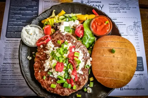 Soczysty kotlet z dodatkami i bułką, serwowany z sosem. Prawdziwa uczta kuchni bałkańskiej.