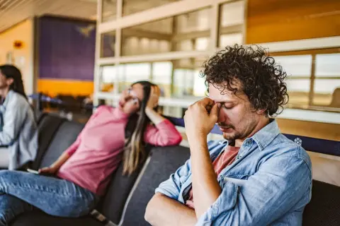 Erschöpfter Mann im Wartebereich, der sich die Augen reibt. Die Frau neben ihm wirkt ebenfalls müde. Gute **Jetlag Tipps** sind hier gefragt.