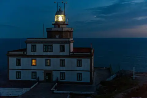 Faro de Finisterre iluminado al anochecer, con el mar oscuro de fondo. Una luz cálida brilla en una ventana.